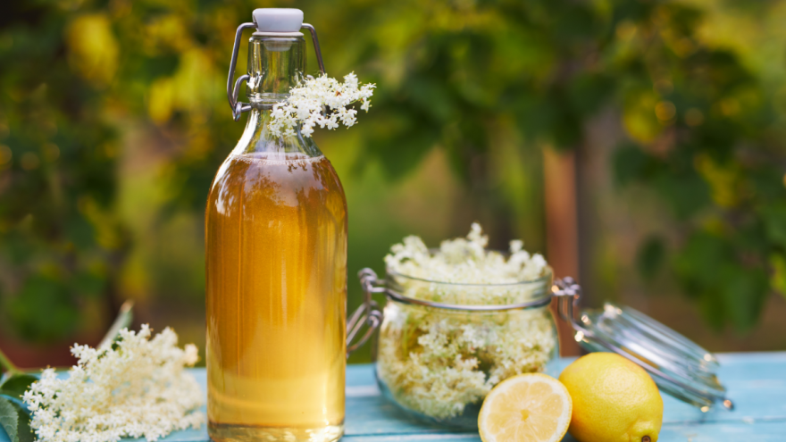 Eine Flasche mit Holunderblütensirup steht neben Blüten und Zitronen auf einem Tisch.