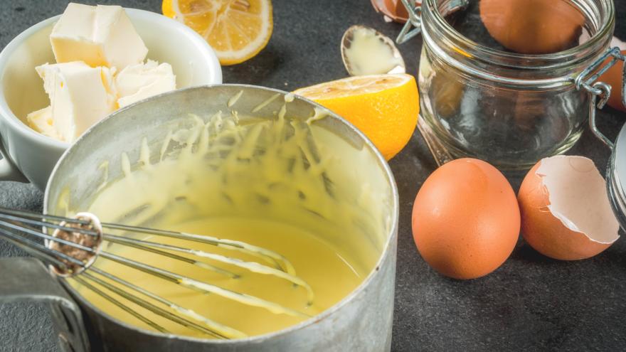 Ein Topf mit Sauce Hollandaise neben Eierschalen, einer Zitrone und Butter