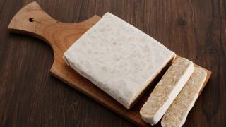 Eine Stück Tempeh liegt auch einem Holzbrett, zwei dünne Scheiben sind abgeschnitten.