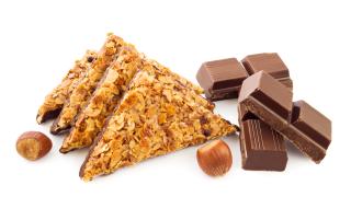 Vier Nussecken, die mit Mandelsplittern bedeckt sind. Daneben Stücke von Schokolade und zwei Haselnüsse