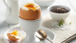 Ein aufgeschlagenes Ei mit sichtbarem Eigelb auf einem weißen Teller, daneben eine kleine Schale mit Gewürzen. Im Hintergrund stehen eine Kaffeetasse, ein Glas Wasser und weiteres Geschirr auf dem Tisch.